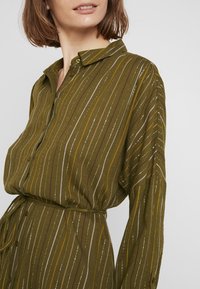 Robe chemise à manches longues verte avec des rayures verticales jaunes, brunes et argentées. Elle présente un col classique et une fermeture à boutons.