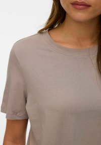 Γυναίκα που φοράει μονόχρωμο μπεζ κοντομάνικο μπλουζάκι με στρογγυλή λαιμόκοψη, εμφανίζοντας το πάνω μέρος του κορμού και μέρος του προσώπου με φυσικά χείλη και καστανά μαλλιά.