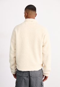 Giacca in fleece crema con superficie testurizzata, colletto alto e maniche lunghe. Presenta una vestibilità comoda e un design leggermente corto.
