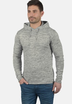Sudadera gris hecha de un tejido suave y texturizado. Con bolsillo frontal, capucha con cordón y diseño ceñido. Combinada con jeans azules.
