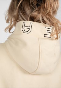 Hoodie color crema con un grande cappuccio. Presenta lettere nere ricamate sul colletto. Realizzato in un tessuto morbido con una texture liscia.