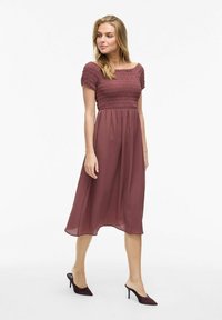Robe midi mauve avec un corsage smocké et des manches courtes, jupe fluide, associée à des talons violets foncés. Tissu léger et semi-transparent.