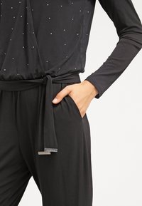 Combinaison noire à manches longues, dotée d'un design croisé, d'accents en pierres, et d'une taille ceinturée ; comprend des poches latérales et une étiquette de logo.