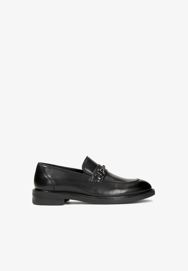 Schwarze Lederloafers mit glatter Textur, runder Spitze und einem dekorativen Metallverschlussakzent auf dem Blatt. Niedriger, gestapelter Absatz.