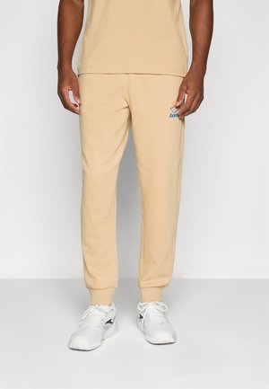 Beige Sweatpants mit elastischem Bund und Bündchen an den Knöcheln, mit einem blauen Logo-Detail. Der Stoff wirkt weich mit einer glatten Textur.