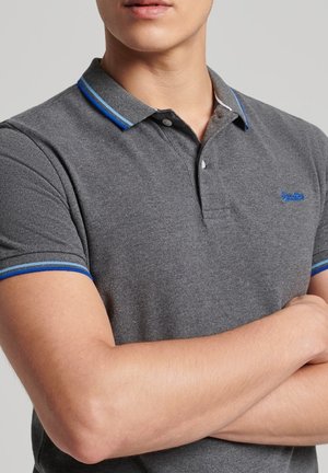 Muž se založenýma rukama, oblečený v šedém polotriku s límečkem a manžetami s modrými pruhy, stojící před jednobarevným pozadím.