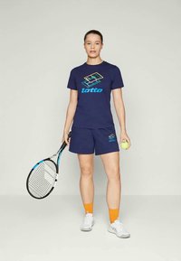 Námořnické sportovní tričko a šortky s logem Lotto, doplněné o oranžové ponožky a bílé tenisky. Drží tenisovou raketu a míček.