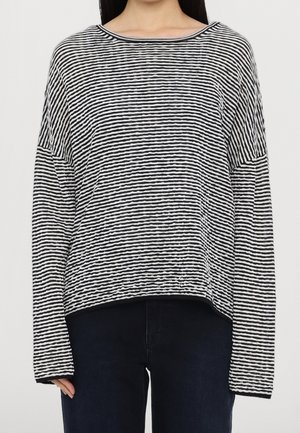 Femme portant un haut à manches longues noir et blanc à rayures horizontales, coupe ample, et un pantalon foncé sur fond uni.