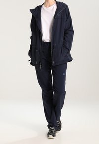 Person wearing navy blue jacket and pants with white t-shirt and black sneakers, standing with hands in jacket pockets against white background.