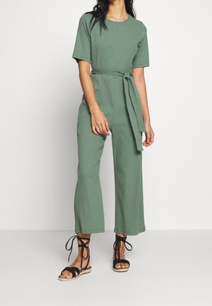 Femme portant une combinaison côtelée verte à manches courtes avec une ceinture à la taille et des sandales plates noires à lacets, debout devant un fond uni.