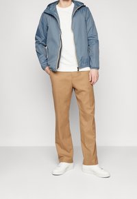 Hellblaue Kapuzenjacke, mitReißverschluss, elastische Bündchen; weißes Sweatshirt darunter; beigefarbene, locker sitzende Hose; weiße Sneakers.