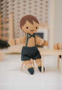 Knuffel met een bruine stoffen lijf, draagt donkere korte broek en groene bretels; heeft een gestikte glimlach en bruine haaraccenten.