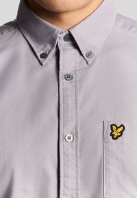 Uomo che indossa una camicia button-down di un leggero colore viola con colletto abbottonato e un piccolo uccellino giallo ricamato sul taschino sinistro.