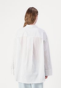Long-sleeve white shirt with light gray stripes, a pointed collar, and buttoned cuffs, featuring a loose fit and back pleat detail.