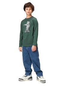 Vihreä pitkähihainen t-paita, jossa on graafinen kuva hupparia käyttävästä karhusta, yhdistettynä löysiin sinisiin farkkuihin ja valkoisiin tennareihin, joissa on mustia yksityiskohtia.