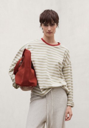 Persoon die een beige gestreept shirt met lange mouwen en een lichte broek draagt en een rode getextureerde handtas vasthoudt tegen een effen lichte achtergrond.