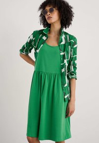Groene katoenen jurk in een mix met een uitlopende rok, gecombineerd met een groen-wit gestreepte blouse. Opvallende details zijn de ronde zonnebril.