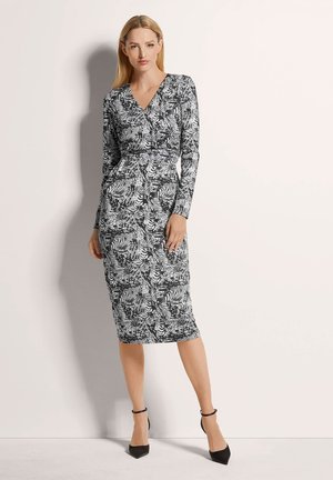 Zwart-witte jurk met lange mouwen en een wikkelontwerp, voorzien van een bladerpatroon, getailleerde silhouet en midi-lengte. Zwarte enkelschoenen met strap.