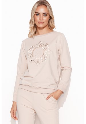 Jasnobeżowy sweatshirt z długimi rękawami, okrągłym dekoltem oraz haftowanym wzorem z literą "M" na środku. Miękka, teksturowana tkanina.