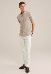Giovane uomo in piedi, che indossa una camicia beige a maniche corte con bottoni, pantaloni bianchi, mocassini marrone scuro e un orologio da polso.