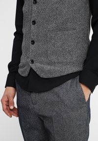 Hombre con chaleco gris abotonado sobre camisa negra y pantalones texturizados grises, con una mano en el bolsillo, sobre un fondo liso.