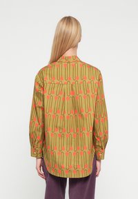 Πουκάμισο με μακριά μανίκια σε ελαιώδες πράσινο, με επαναλαμβανόμενο μοτίβο από ροζ τουλίπες και γραμμές. Μανσέτες με κουμπιά και ένα πτυχό στο πίσω μέρος.