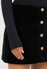 Melnā samta A-formas svārki ar zeltītām pogām, gluda tekstūra un pieguļoša josta, uz kuras atpūtina roku.