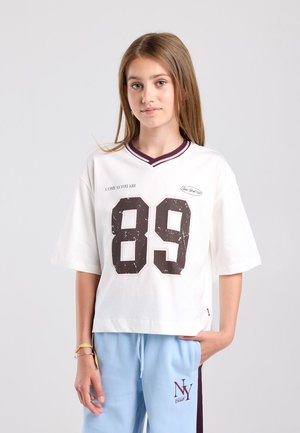 Giovane con i capelli lunghi che indossa una maglietta bianca con il numero 89 e pantaloni azzurri con il logo NY Club, su sfondo uniforme.