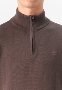 Rjav zip-up pulover z rebrastim ovratnikom in majhnim logotipom na prsih. Tkanina se zdi mehka z subtilno teksturo.