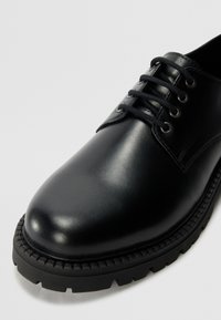 Zapato de cuero negro con superficie lisa, puntera redondeada y suela texturizada en contraste. Presenta cordones y ojales negros para un ajuste seguro.