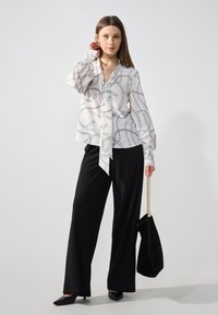 Blusa bianca con motivo a catena nera, scollo a V, lunghe maniche voluminose, legata davanti. Pantaloni neri a gamba larga e una borsa nera con tracolla in catena dorata.