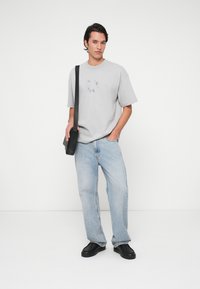 Siva kratki majica s kratkimi rokavi s krožnim grafičnim dizajnom, v kombinaciji s svetlo modrimi ohlapnimi jeans hlačami, črnimi natikači in črno torbo preko ramena.