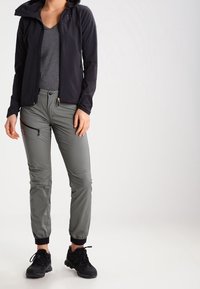 Černá lehká bunda s kapucí, v kombinaci se šedými zúženými kalhotami se zipovými kapsami a černými lemovanými kotníky, nošená s černou obuví.