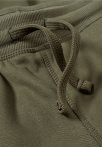 Olivengrønt stof med en blød tekstur, med en snøre-lukning. Linningen har ribstrik og synlige syningsdetaljer.