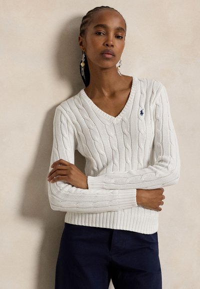 Femme aux cheveux tressés portant un pull blanc à col en V en maille torsadée à manches longues et un pantalon foncé, posant bras croisés devant un fond uni.