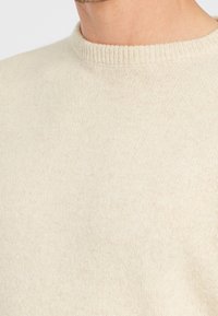 Beige tröja med ribbad halsringning, tillverkad av mjukt, texturerat stickat material, med en klassisk, rak skärning.