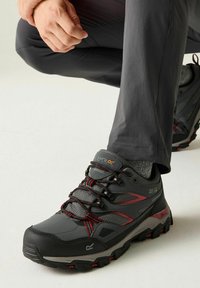 Persona atándose un zapato de senderismo gris oscuro y negro con detalles rojos, llevando calcetines grises y pantalones gris oscuro, sobre un fondo claro.
