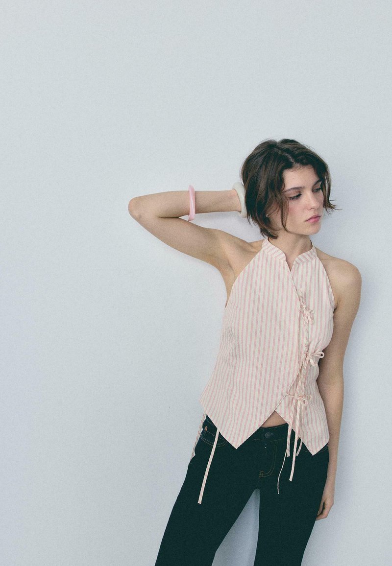 Jeune femme aux cheveux courts portant un haut rayé sans manches et un pantalon foncé, posant avec un bras levé et la main derrière la tête.