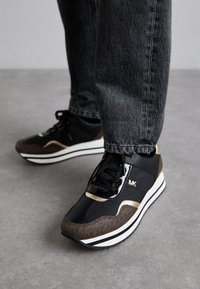 Zwarte en bruine sneakers met een gestructureerde afwerking, voorzien van een gouden accent, een witte tussenzool en zwarte veters, gecombineerd met een paar grijze jeans.