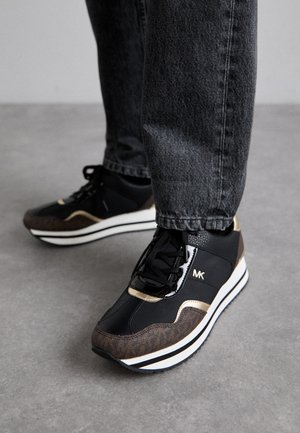 Sneaker neri e marroni con una finitura testurizzata, dotati di un accento dorato, suola intermedia bianca e lacci neri, abbinati a jeans grigi.