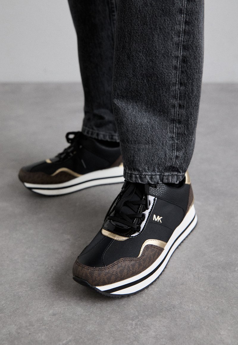 Zwarte en bruine sneakers met een gestructureerde afwerking, voorzien van een gouden accent, een witte tussenzool en zwarte veters, gecombineerd met een paar grijze jeans.