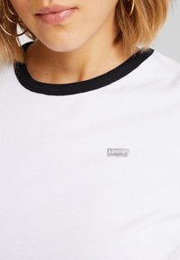 Vit bomull t-shirt med svart ribbad krage och en liten silver Levi's-logotyp på bröstet. Slät textur, avslappnad design.