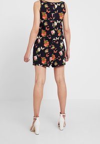 Mono negro de flores que presenta un top sin mangas, cintura elástica y pantalones cortos. Acentuado con flores y mariposas multicolores.