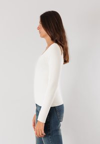 Maglione bianco a coste con maniche lunghe, scollo a V e silhouette aderente, abbinato a jeans di denim blu scuro, visto di lato.