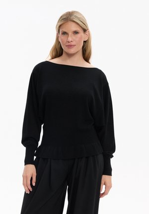 Kobieta o blond włosach, ubrana w luźną czarną bluzkę z długim rękawem i czarne spodnie o szerokich nogawkach, stojąca na białym tle.