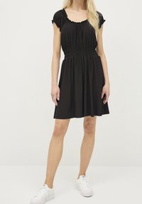 Schwarzes Kleid aus weichem Stoff mit einem gerüschten Bund, kurzen Rüschensäumen und einem lockeren Rock, kombiniert mit weißen Sneakers.