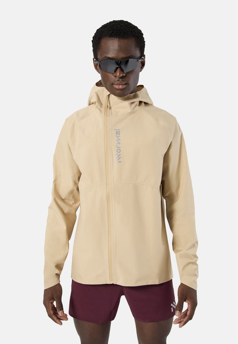 Hombre con chaqueta beige con cremallera y capucha y pantalones cortos de correr granate, con gafas deportivas negras, de pie contra un fondo blanco.