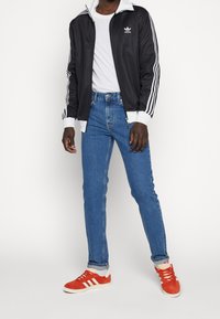 Svart zip-up jacka med vita detaljer och tre ränder, vit t-shirt, blå jeans och röda sneakers med vita ränder. Standard passform.