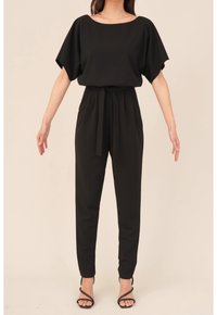 Femme portant une combinaison noire ceinturée avec des manches larges et des jambes fuselées, associée à des sandales à talons hauts noires à lanières.