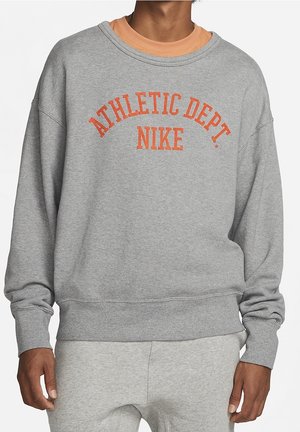 Mężczyzna ubrany w szarą bluzę Nike z napisem „Athletic Dept. NIKE” w pomarańczowym kolorze, w połączeniu z dopasowanymi jasnoszarymi spodniami dresowymi.